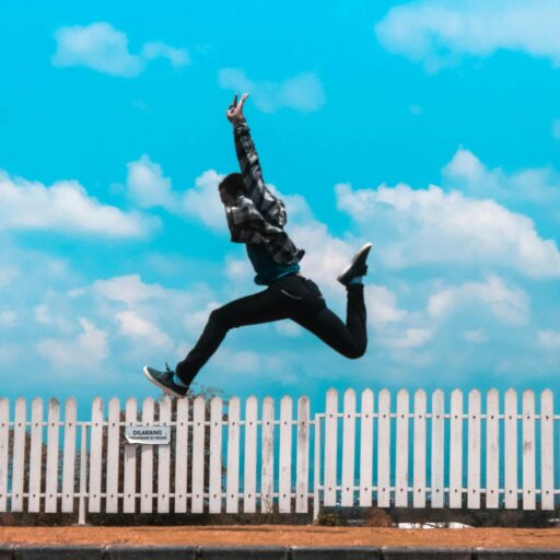 Mid-air capture of a person leaping in front of a picket fence under a bright blue sky.